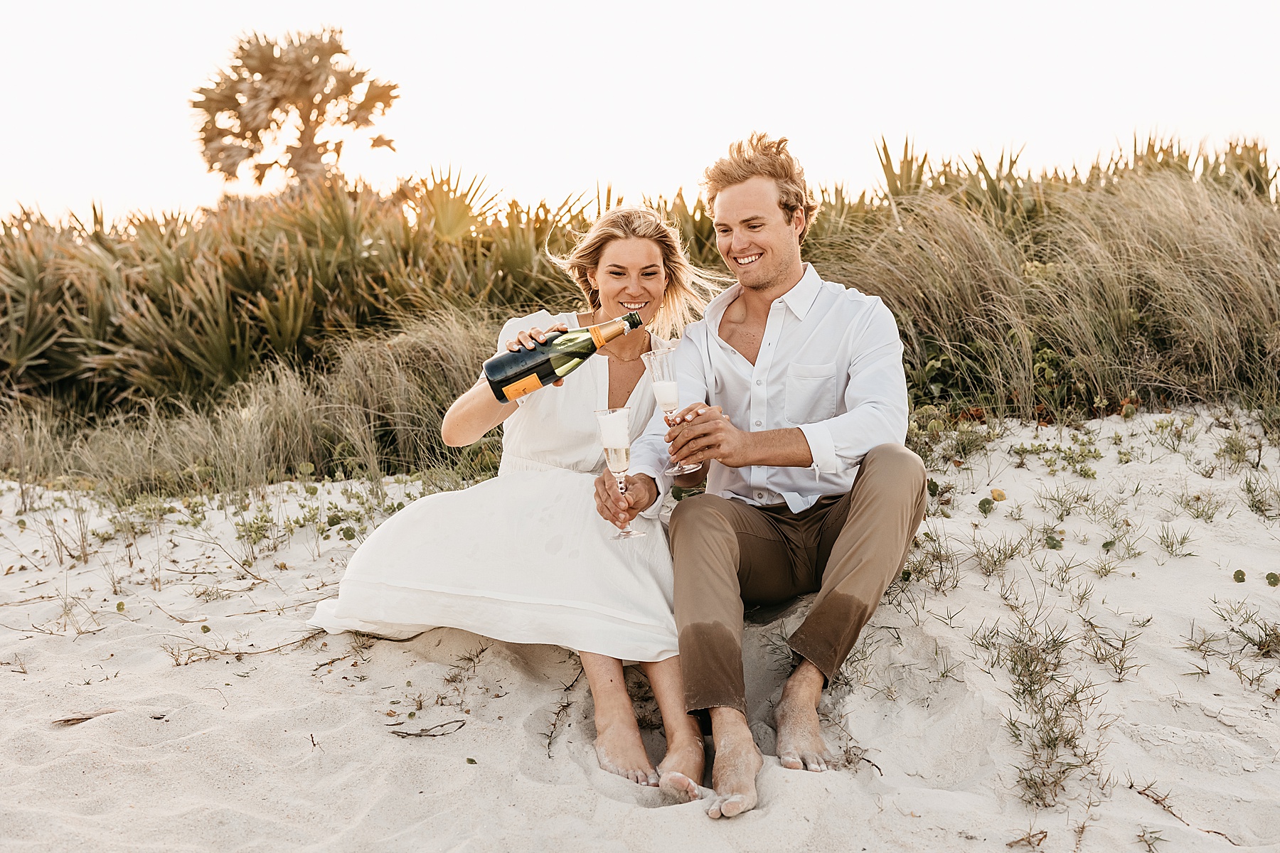 man and woman toasting champagne on the beach at sunset in neutral clothing