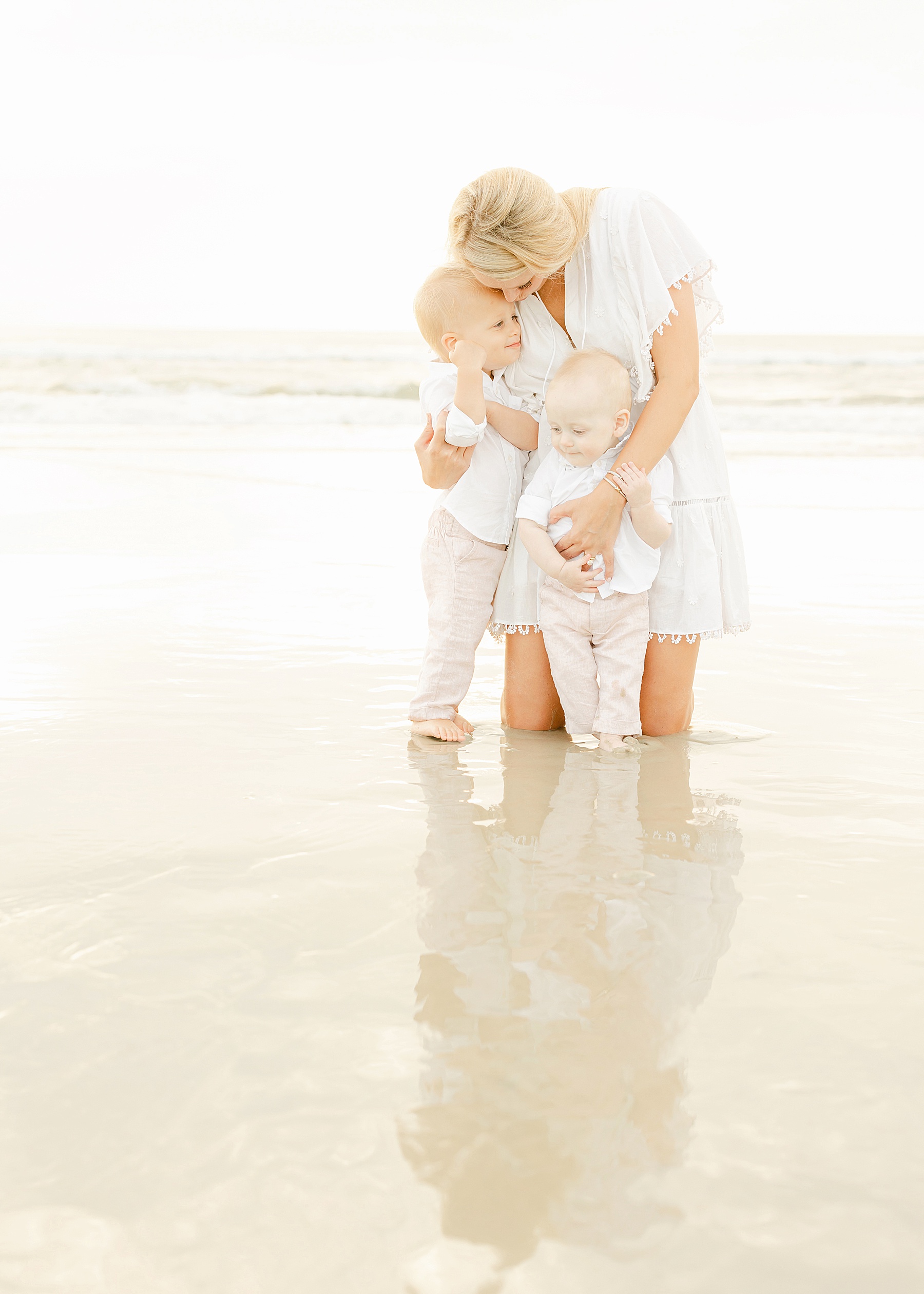 woman holding baby boys on the beach at sunrise wearing white dress in airy and soft lighting