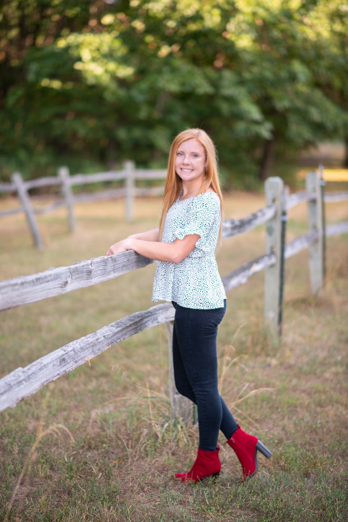 Senior girl in printed top and red boots standing next to a split rail fence in Reeds Spring.