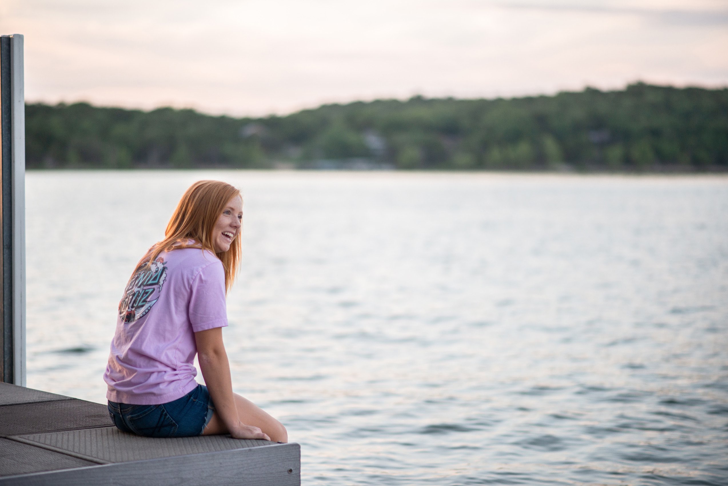 Senior girl in pink tshirt laughing as she sits on the dock at sunset on Tablerock Lake.