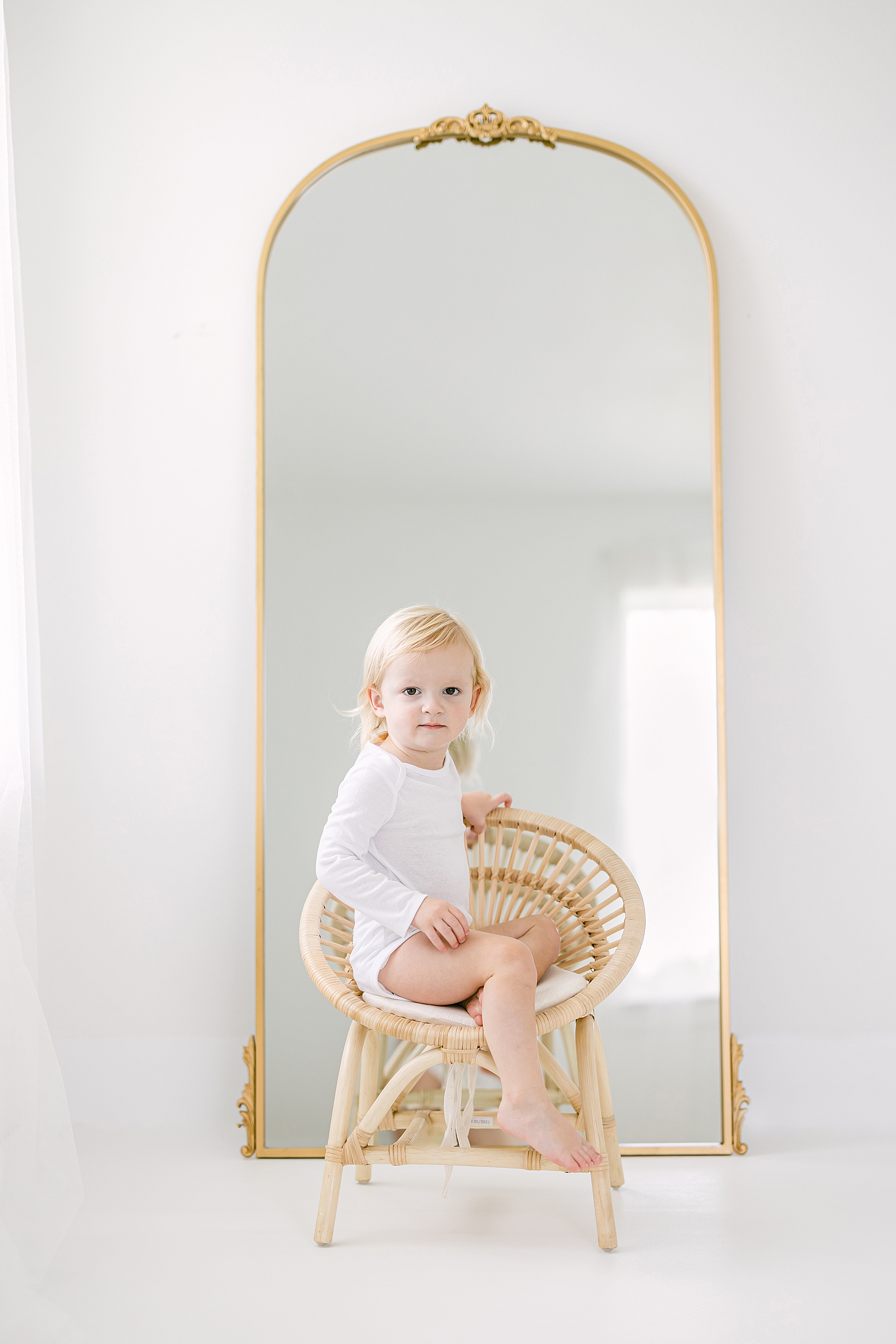 baby boy in white onesie sitting in rattan chair in front of gold floor mirror