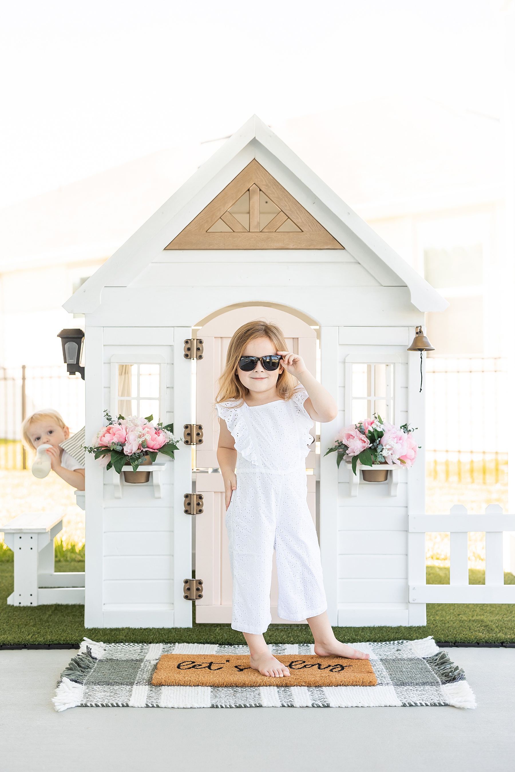 two little kids in white playhouse with pink door laughing at the camera