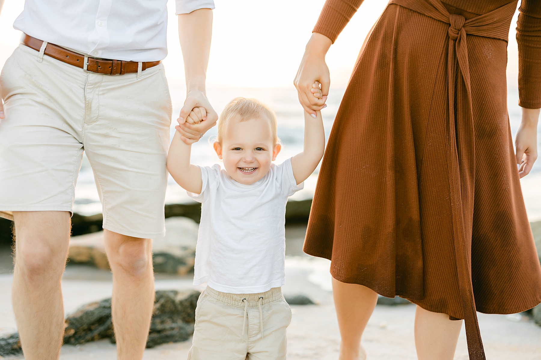 little boy in white shirt and khaki shorts holding his parents hands on the beach at sunrise