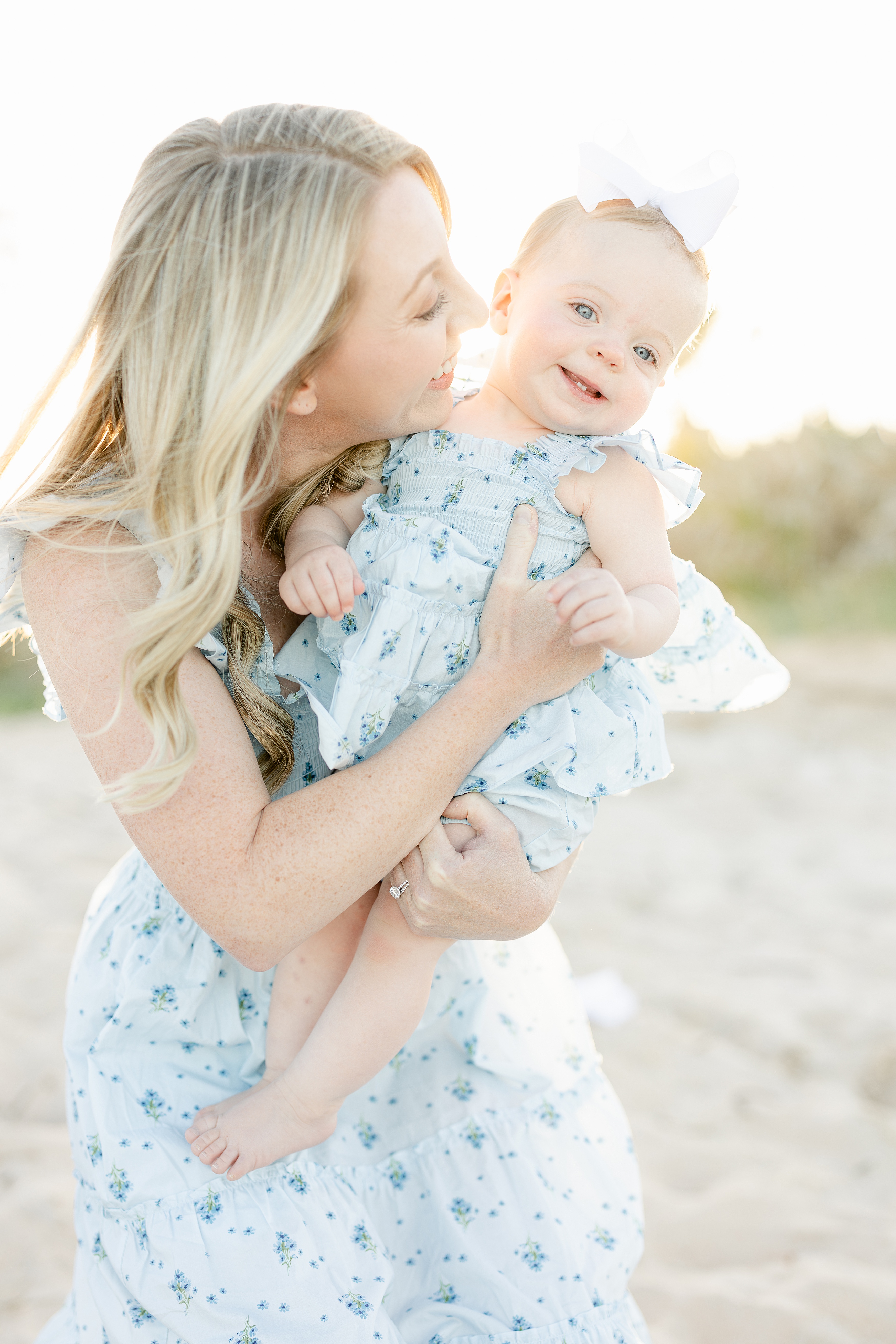 blonde woman in blue and white floral dress holding baby girl on the beach at sunset