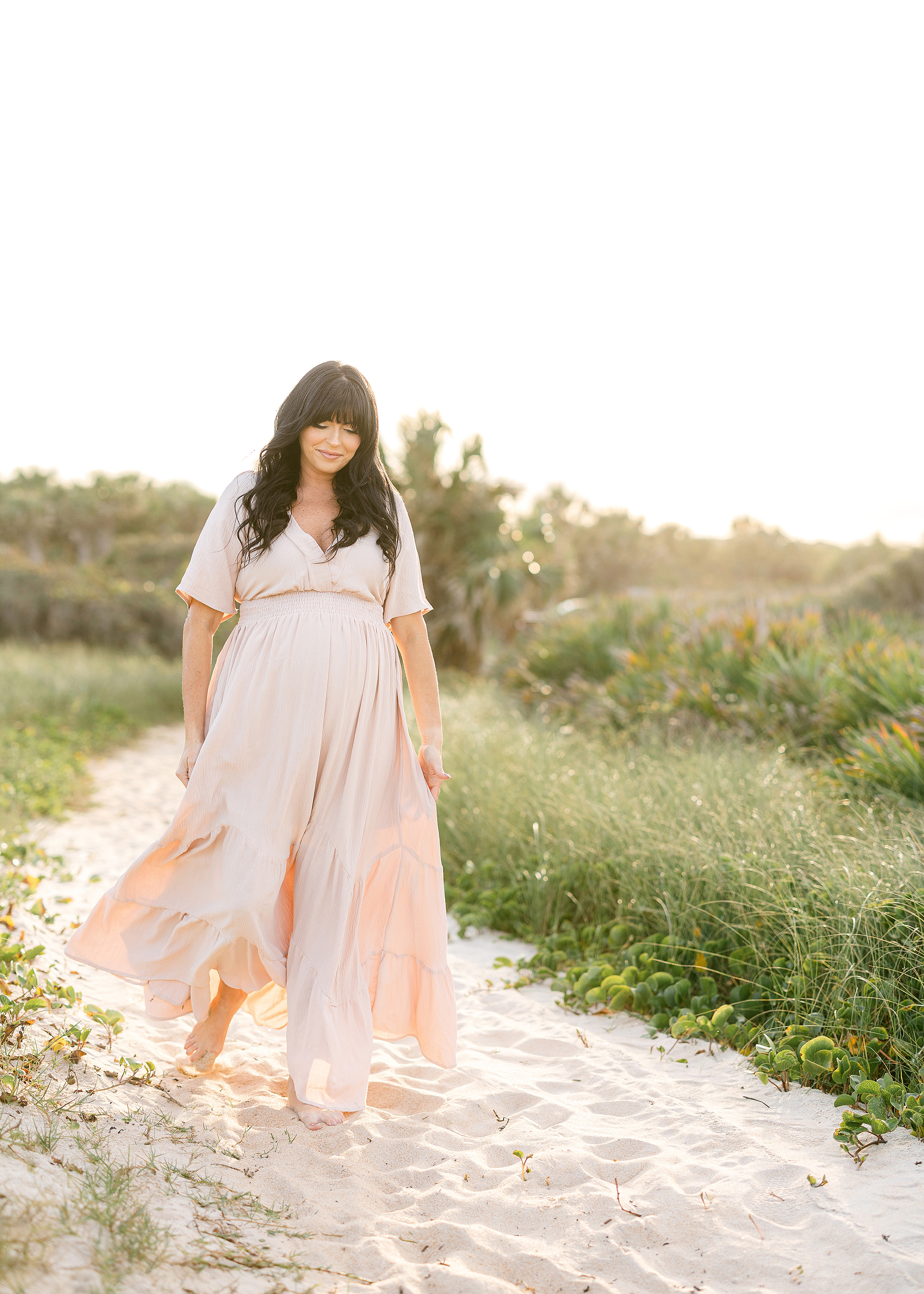 Sun-filled maternity portrait of a woman walking on the beach at sunset.