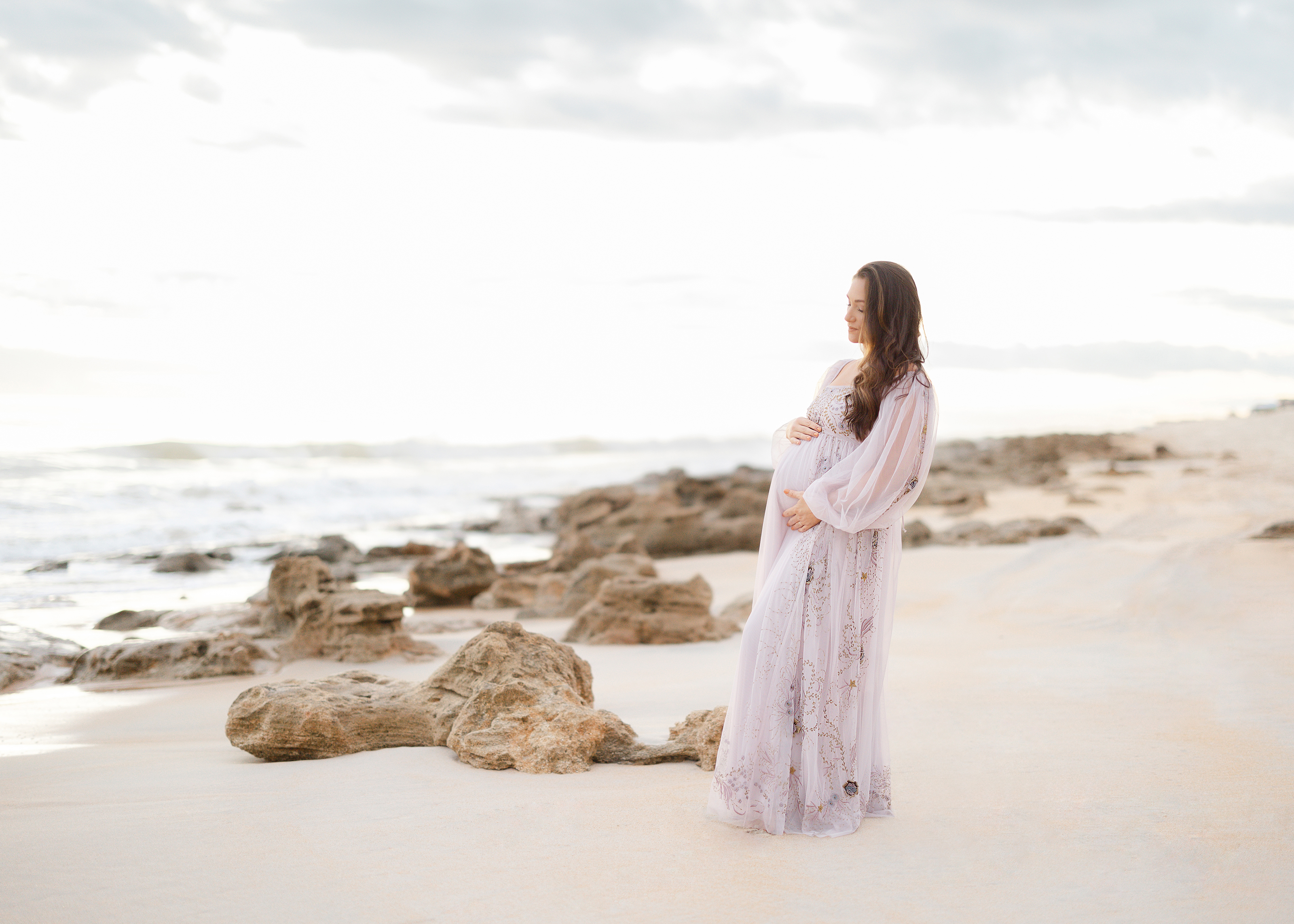 Light and airy sunrise maternity portrait of a woman in a pastel purple dress standing on Saint Augustine Beach.