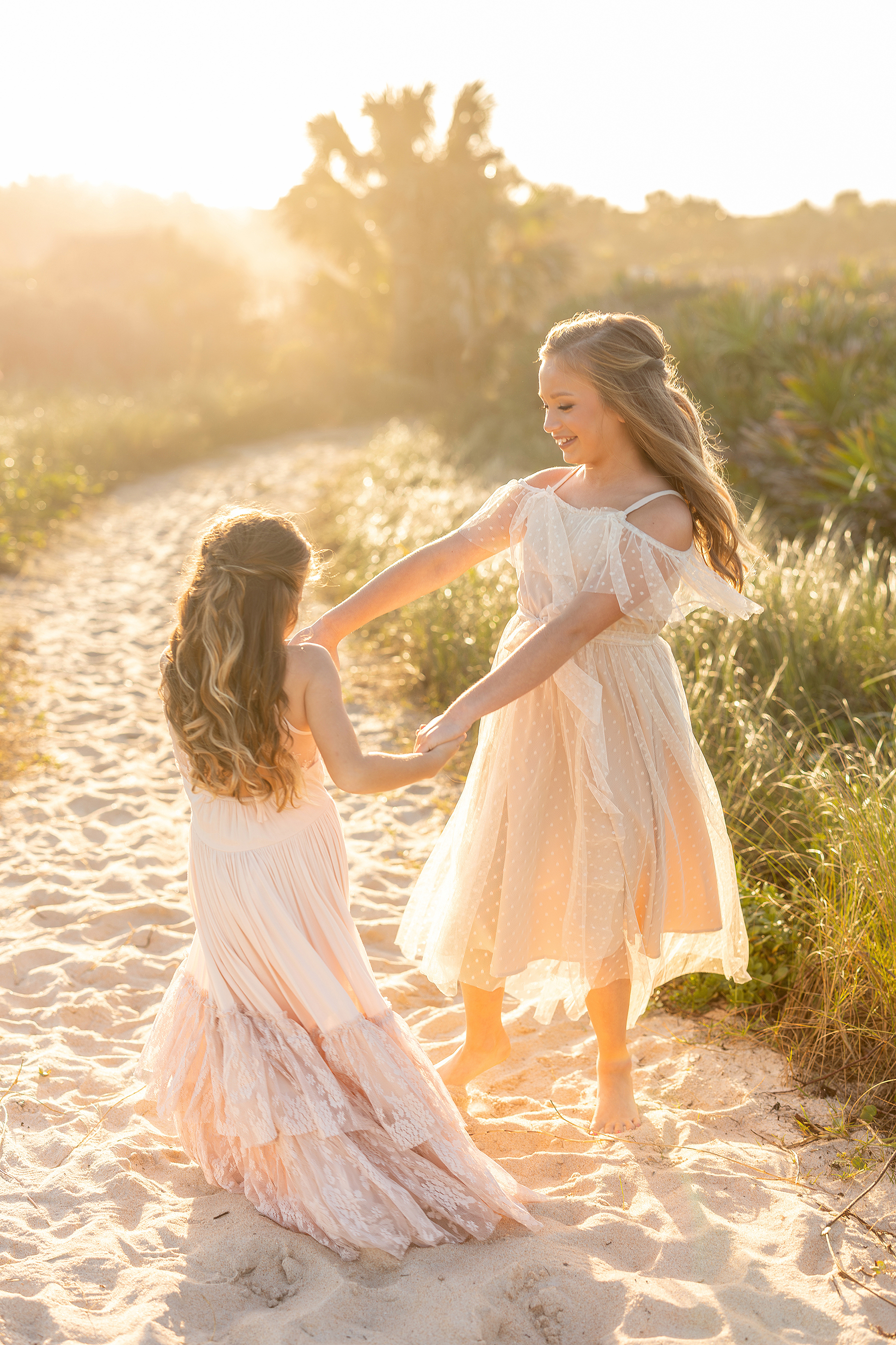 Two little girls in tulle dresses dance together at sunset on a path in the sand.
