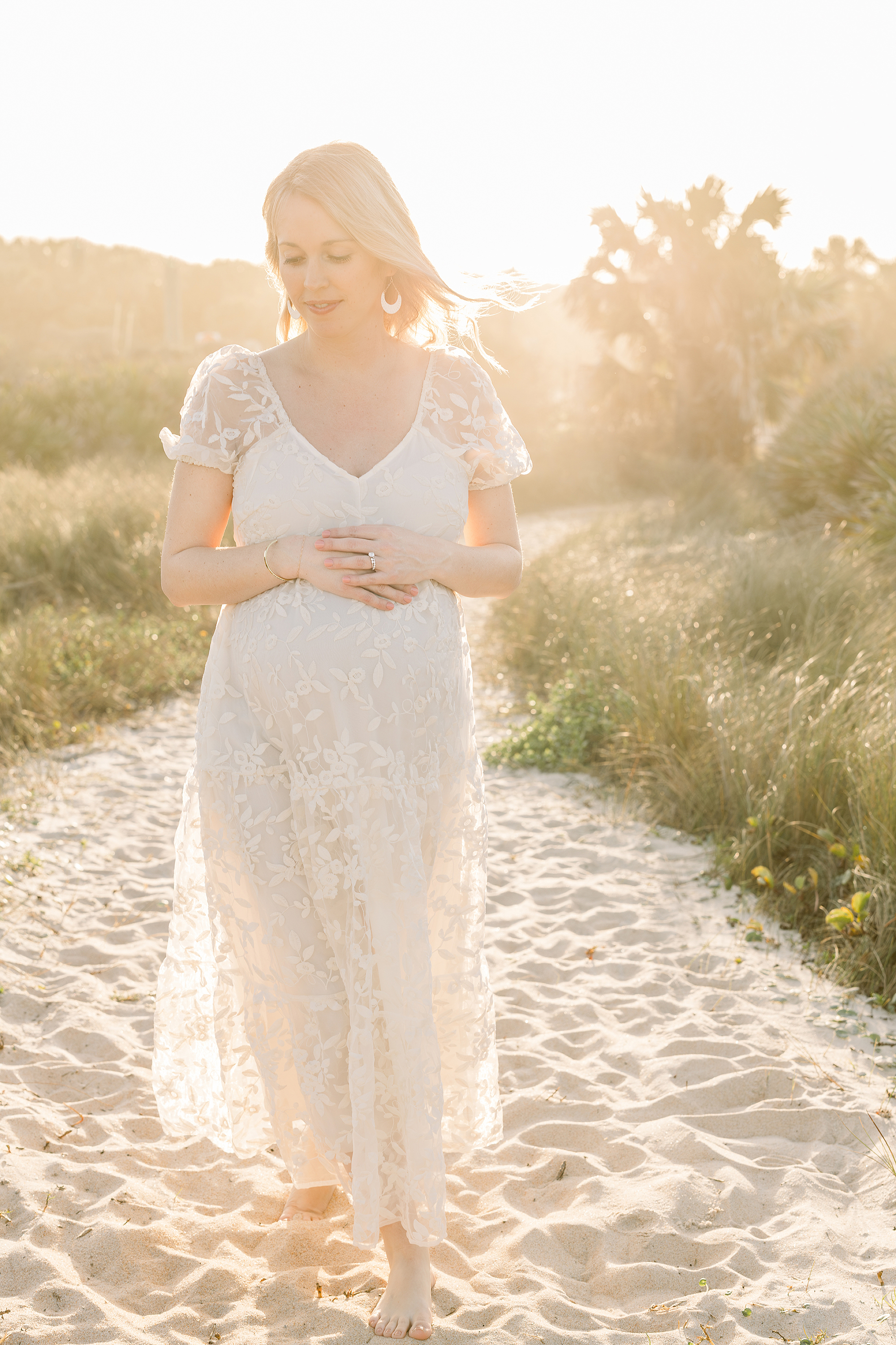 A light filled Golden Hour maternity portrait of a woman in a cream dress standing on the sand in St. Augustine Beach, Florida.