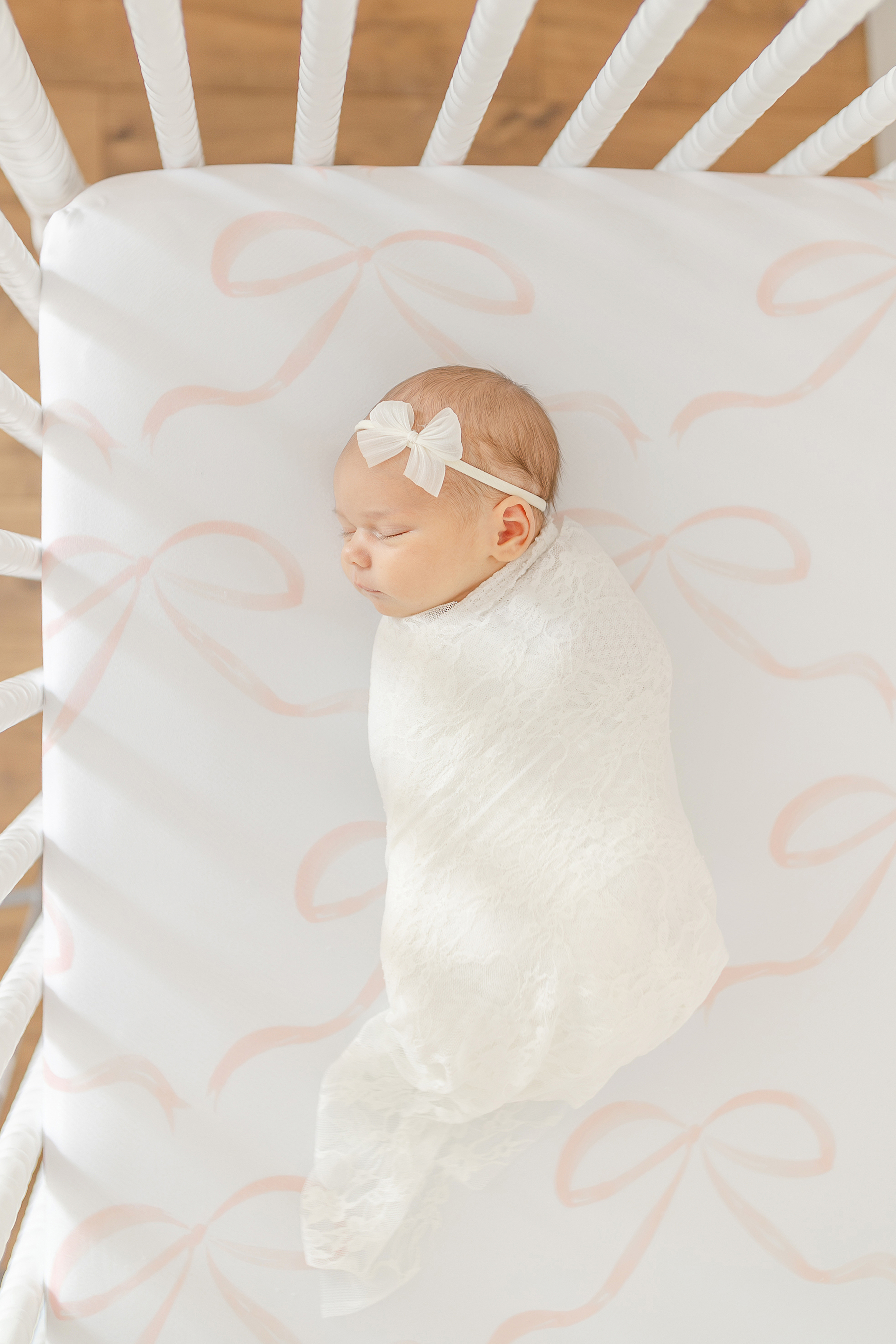 A newborn baby gilr wrapped in a white swaddle in her crib with white sheets with pink bows.