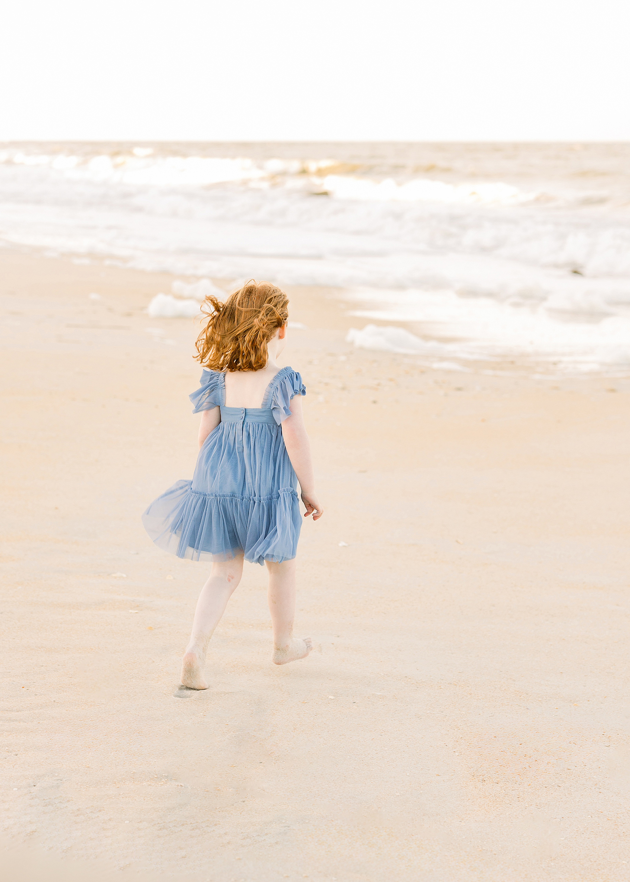 An ethereal childhood beach portrait of a little girl with red hair running towards the water.
