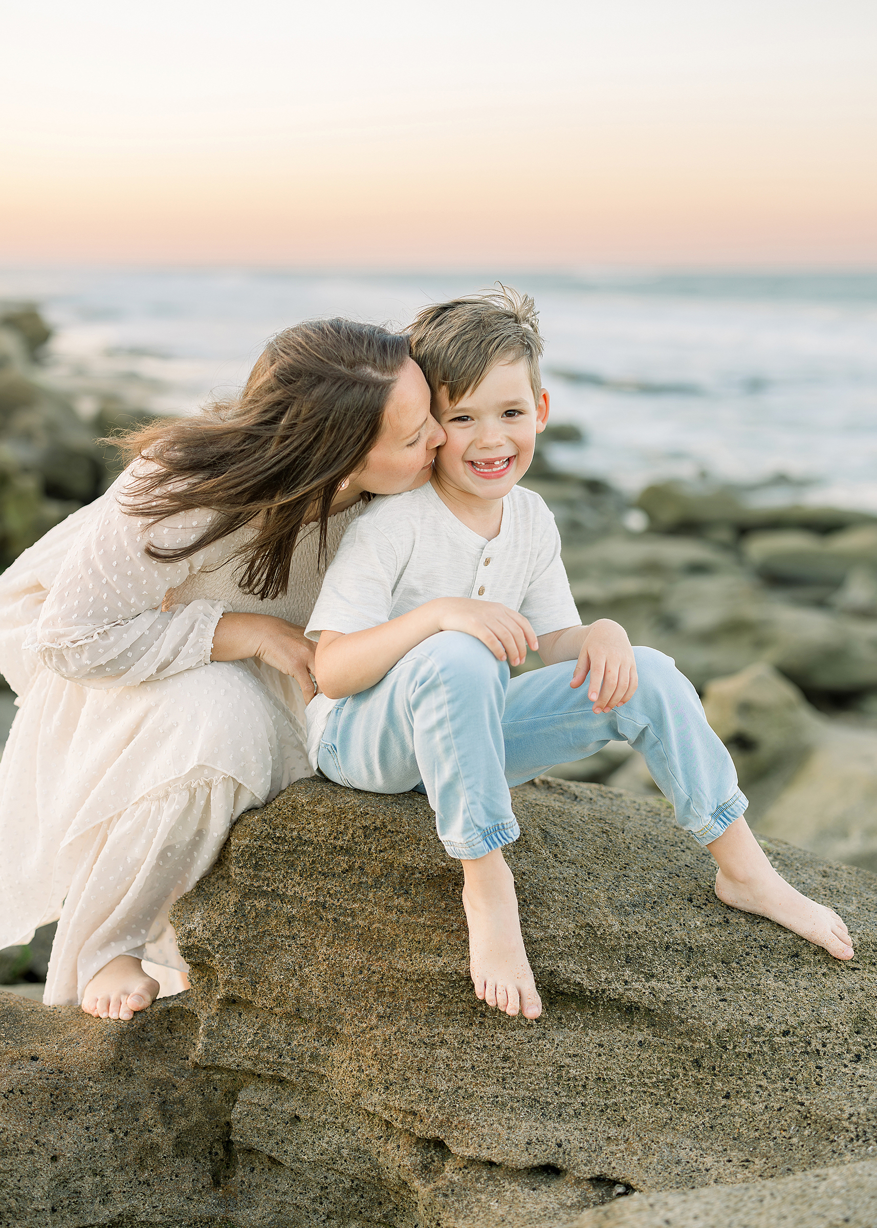 A beach portrait of a mother and her son sitting on the rocks on the beach at sunset.