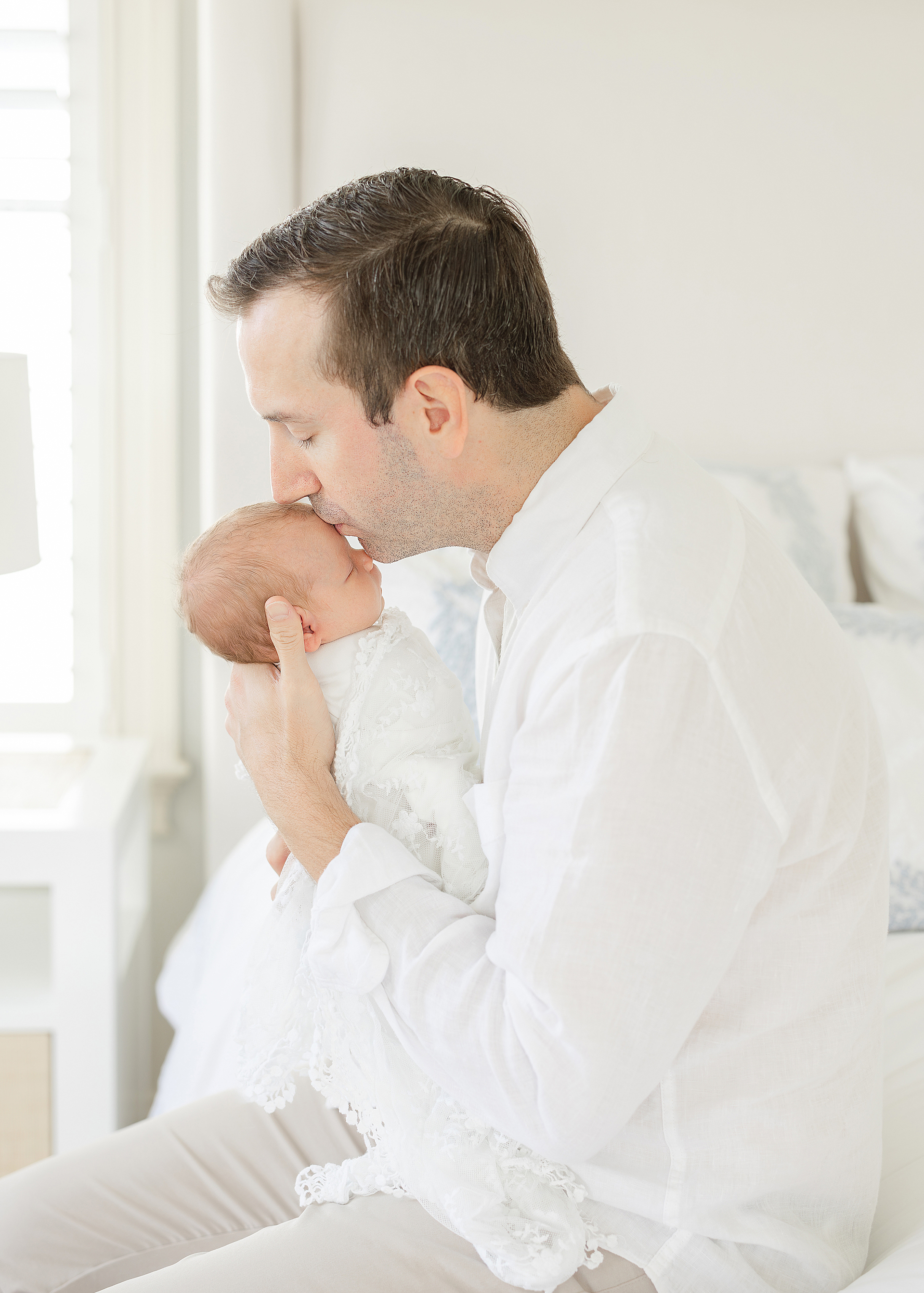 A man holds and kisses his newborn baby girl in front of a white window all dressed in white.