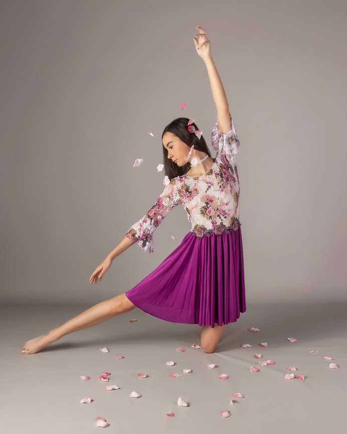 Ballet dancer in a floral top and purple skirt performs gracefully with rose petals scattered on the floor.