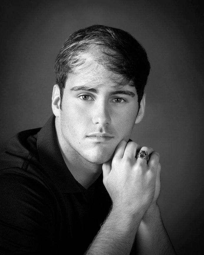 Man in black shirt, contemplatively posing with hands clasped under chin in black-and-white portrait.