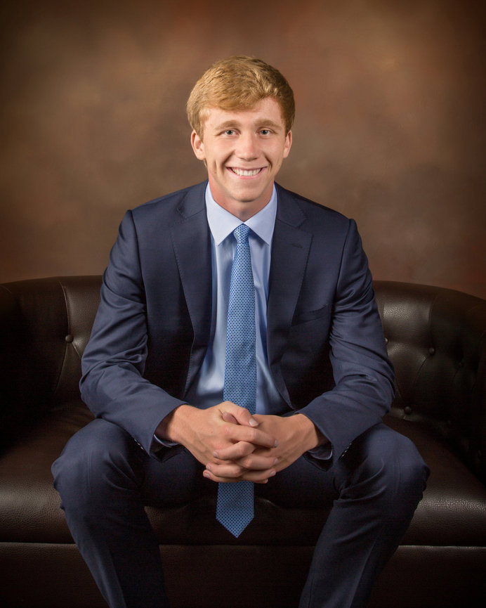 Young man in a blue suit and tie sitting on a leather sofa with a brown background.