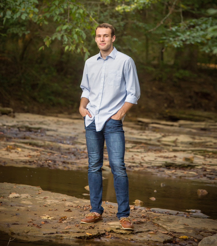 A person in a light blue shirt and jeans stands by a stream in a wooded area.