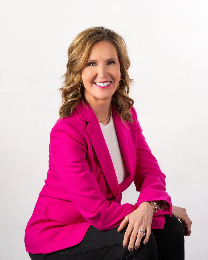 Woman in a bright pink blazer sits and smiles against a plain background.