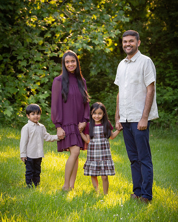 A family of four stands together on grass in front of green foliage, smiling for a portrait.