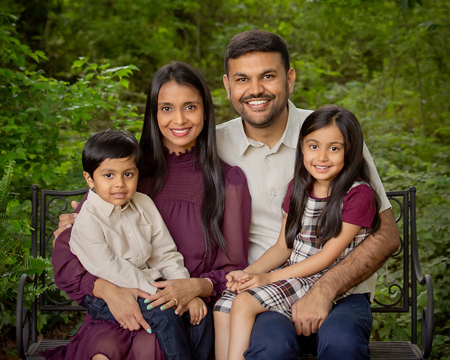A family of four sits on a bench in a garden, smiling at the camera.