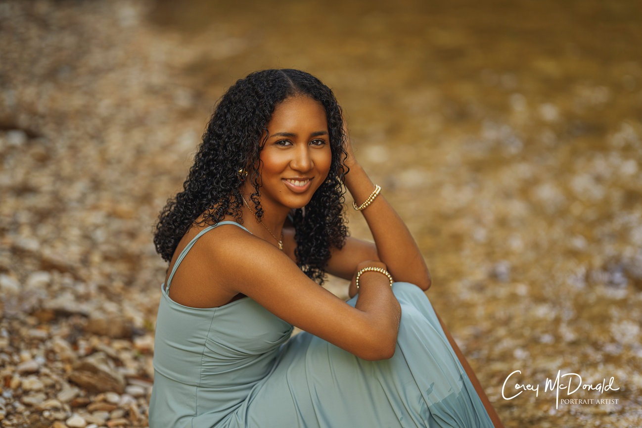 A woman with curly hair sits by a rocky shoreline wearing a sleeveless dress, smiling.