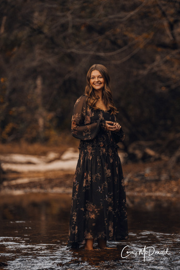Woman in a long floral dress stands smiling in a shallow creek surrounded by trees.