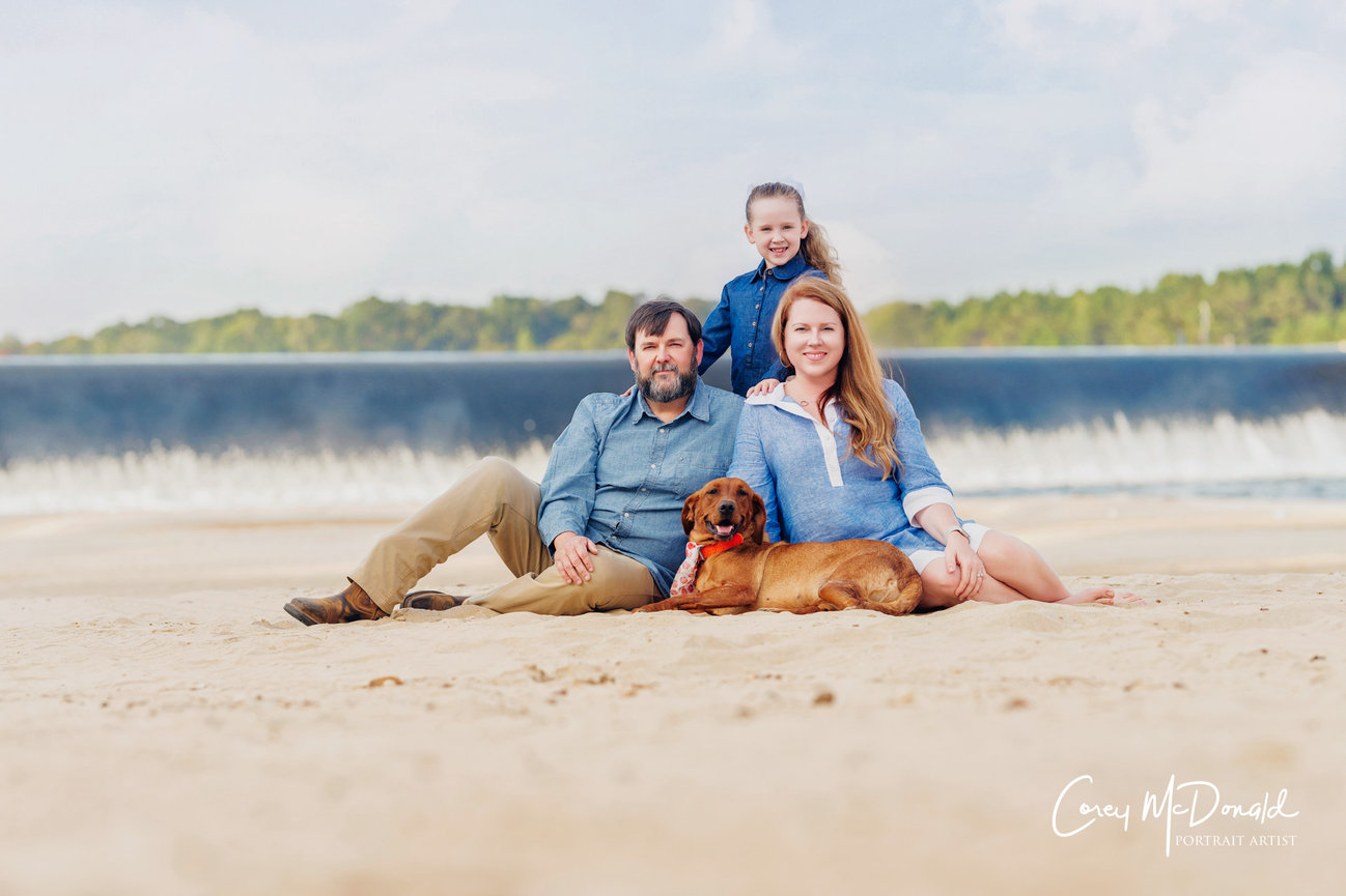 Family sitting on a sandy beach with a dog, a waterfall and trees in the background.