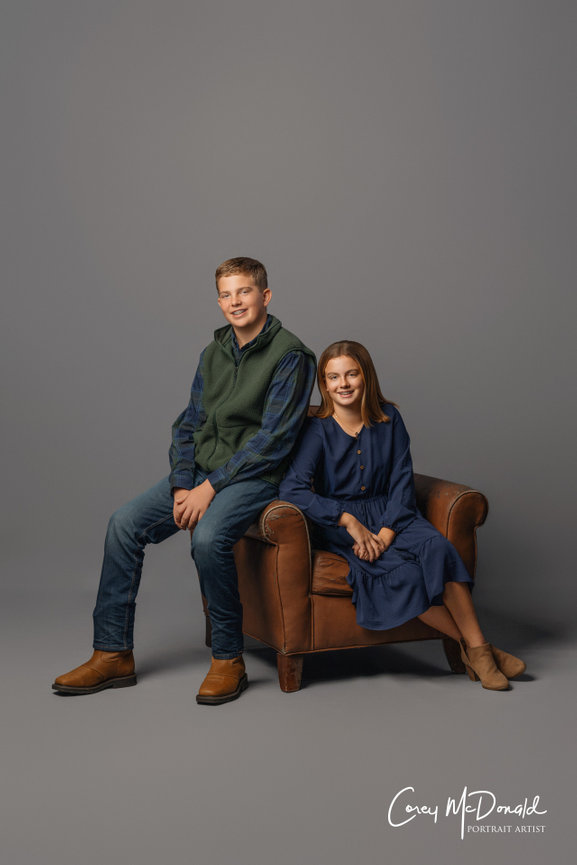 A teenage boy sitting on the back of a brown armchair and a teenage girl seated in it, both smiling.