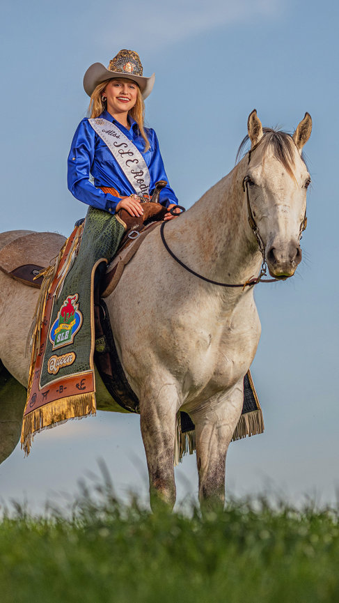 barrel racing girl in rodeo attire and rodeo crown sitting on horse outdoors with blue skies and green grass