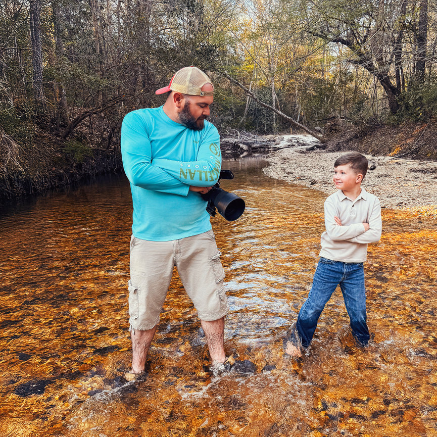 Man and boy standing in a shallow creek, playfully facing each other, surrounded by trees.