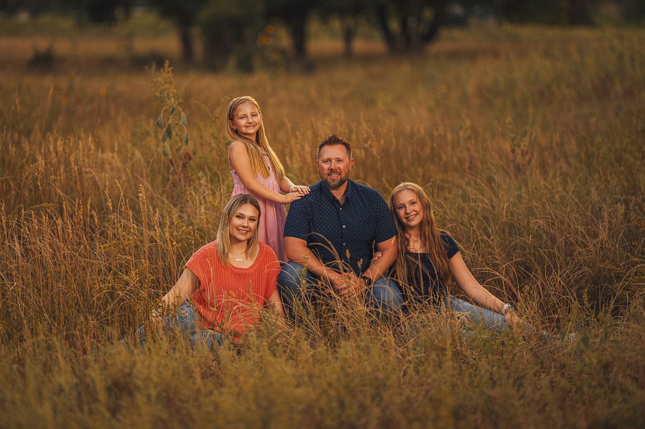 Family of four sitting in a field with tall grass, wearing casual clothes, and smiling at the camera.