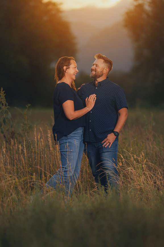A couple smiling at each other, standing in a grassy field at sunset.