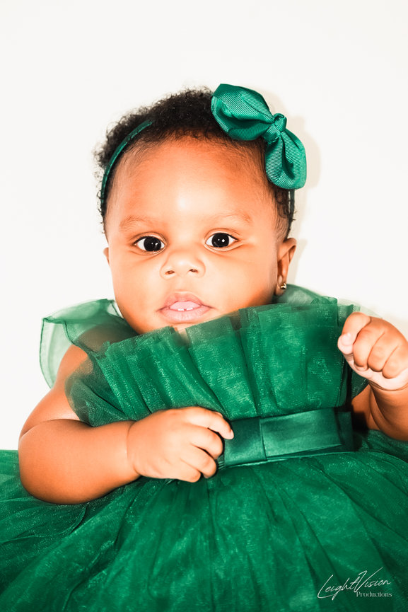 A joyful baby girl dressed in a green dress with a matching bow, radiating innocence and charm.