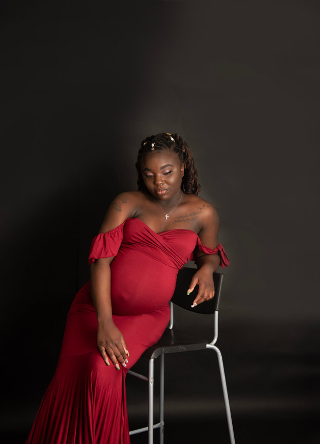 Pregnant woman in a red off-shoulder dress posing on a chair against a black background.