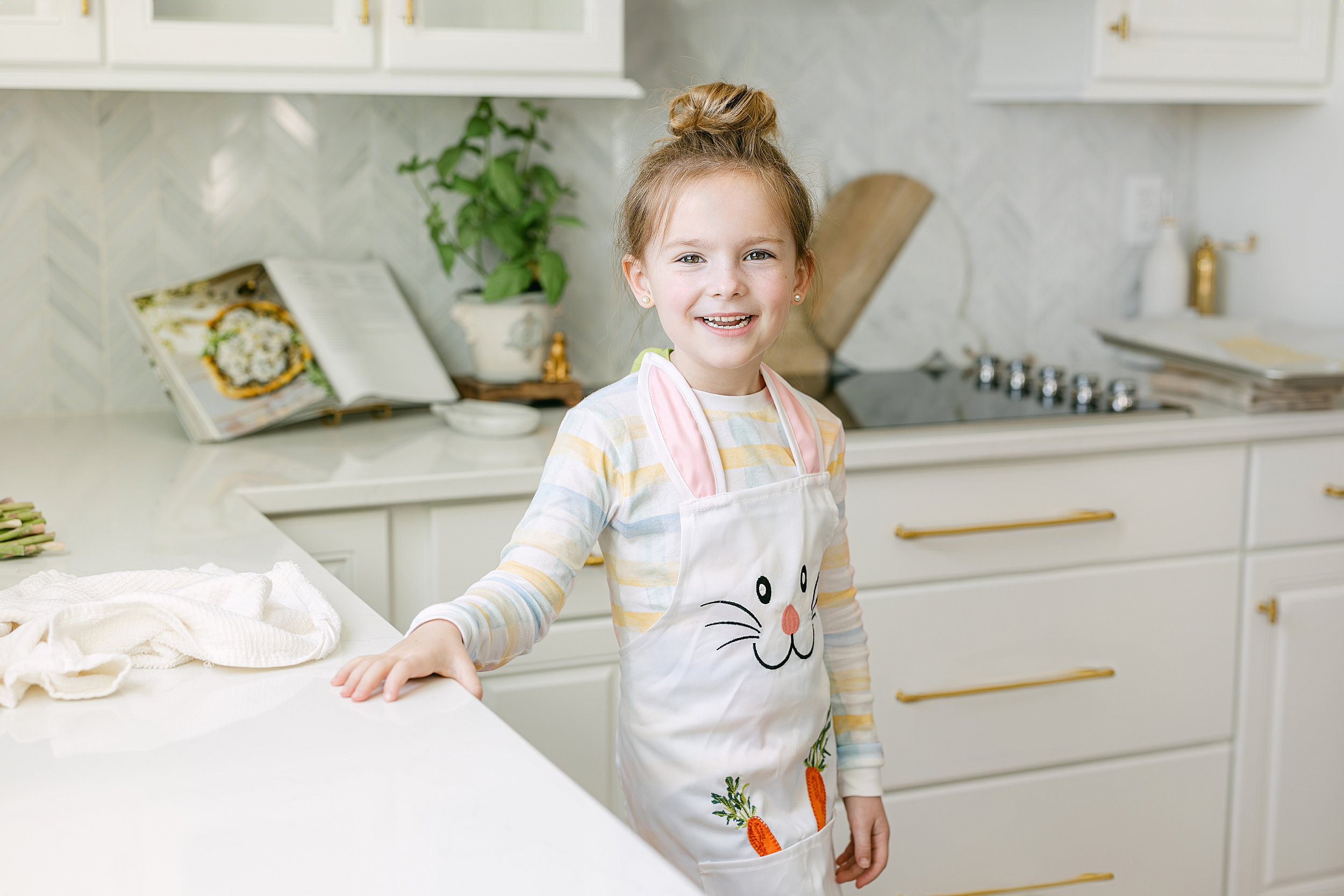 A little girl wearing pastel pajamas and a bunny apron stands in a white kitchen on Easter Sunday.