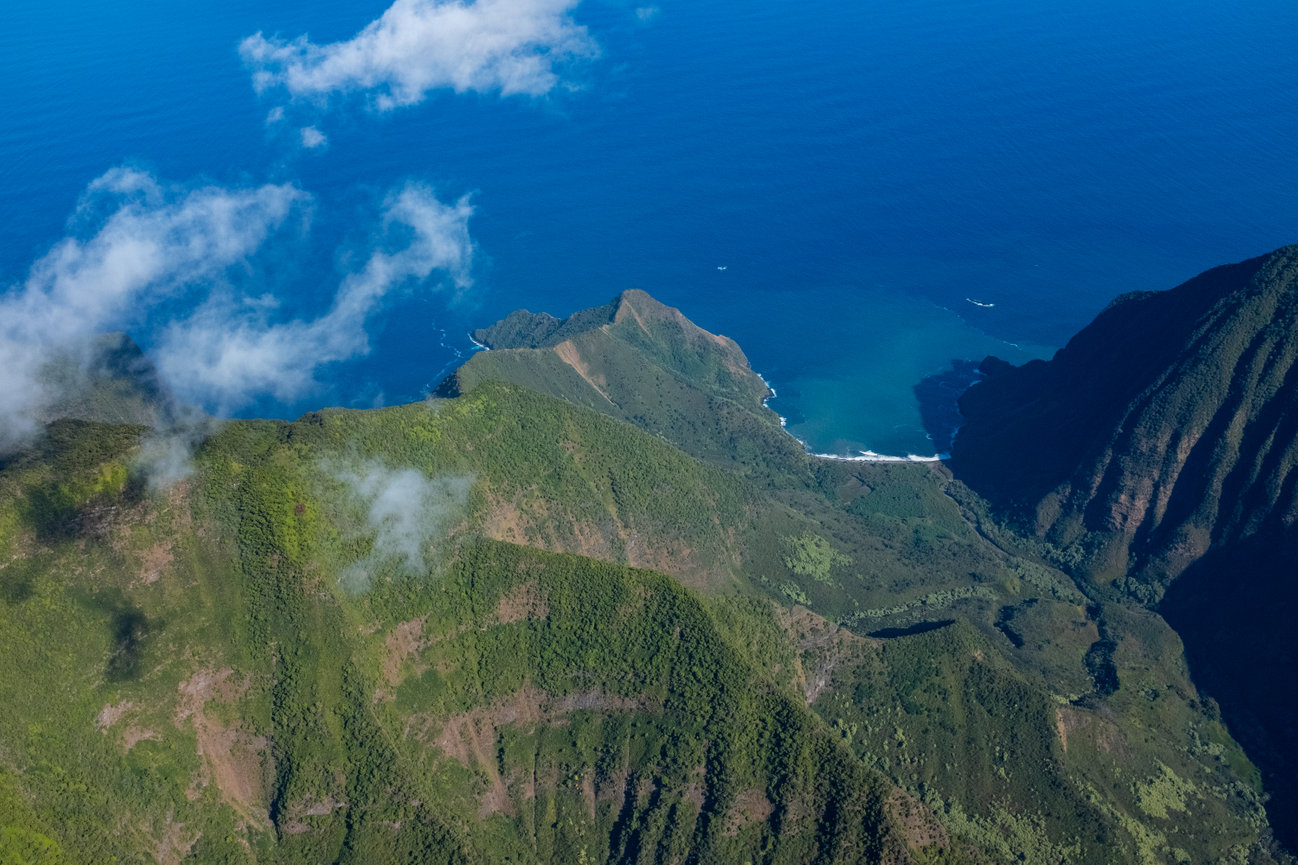 Green jagged cliffs of Molokai from above with wispy clouds and blue ocean in the background.