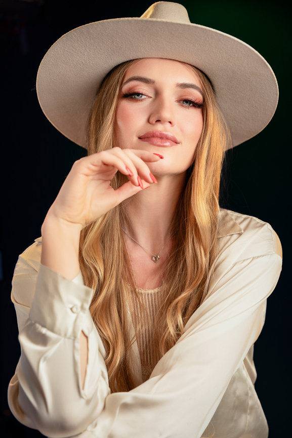 Woman in a beige hat and blouse, with long blond hair, posing confidently against a dark background.