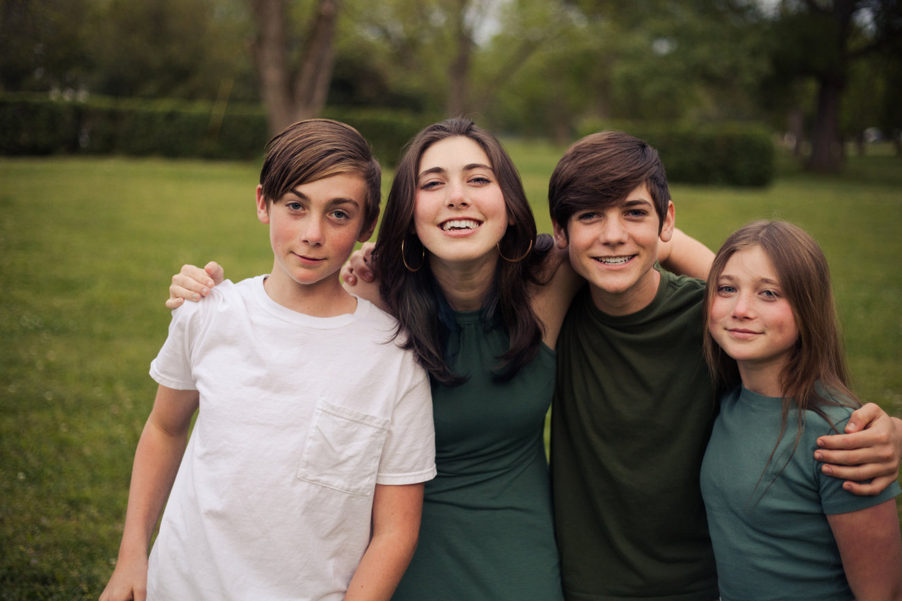 Four siblings standing close together and smiling in a park.