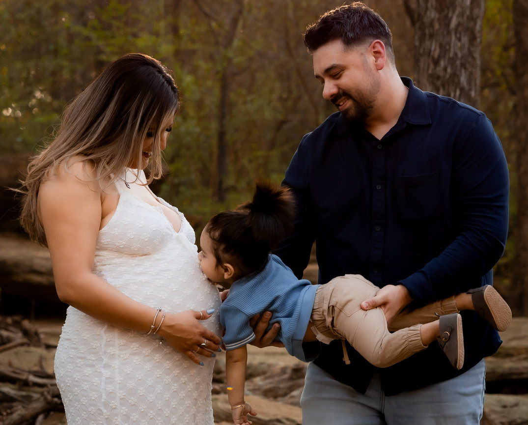 A family in a forest, with a child playfully interacting with a pregnant woman’s belly.
