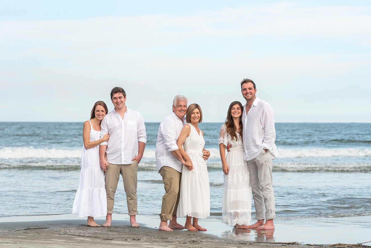 Family posing barefoot on a beach, dressed in white outfits, with ocean waves in the background.