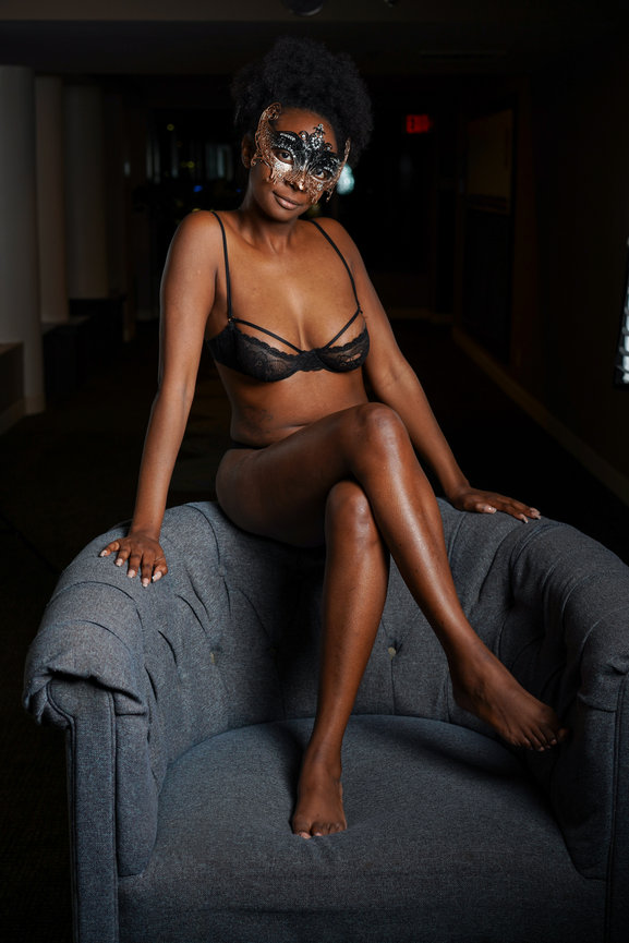 A woman wearing a lace mask and lingerie sits elegantly on a gray chair in a softly lit environment.
