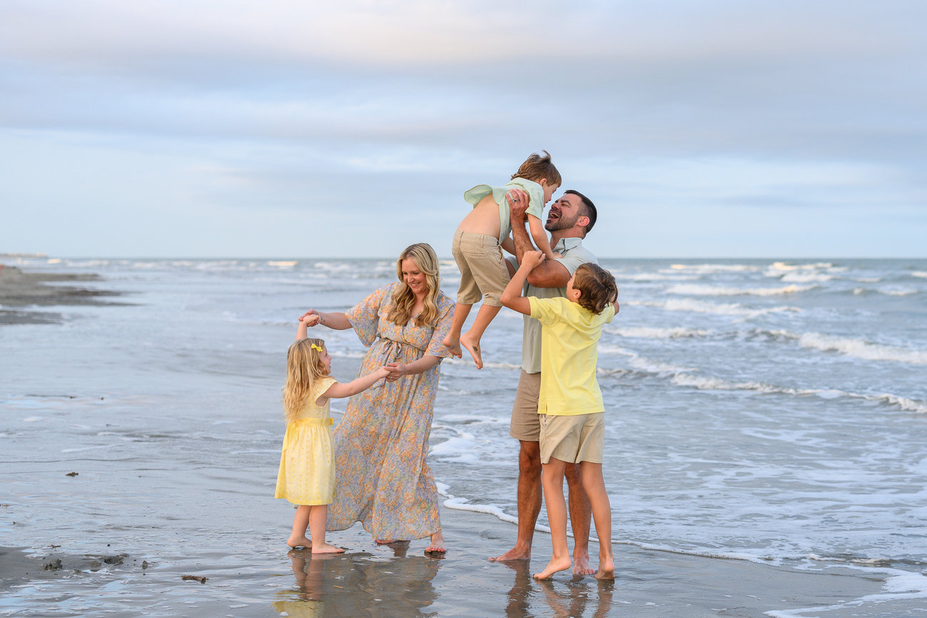 Family enjoying a playful moment on a sandy beach, with waves in the background under a partly cloudy sky.