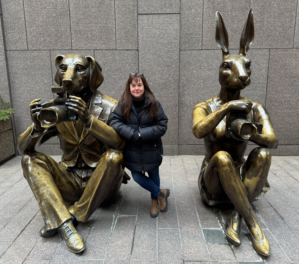 Photographer stands between two statues: a dog with a camera and a rabbit with a camera, both seated outside.