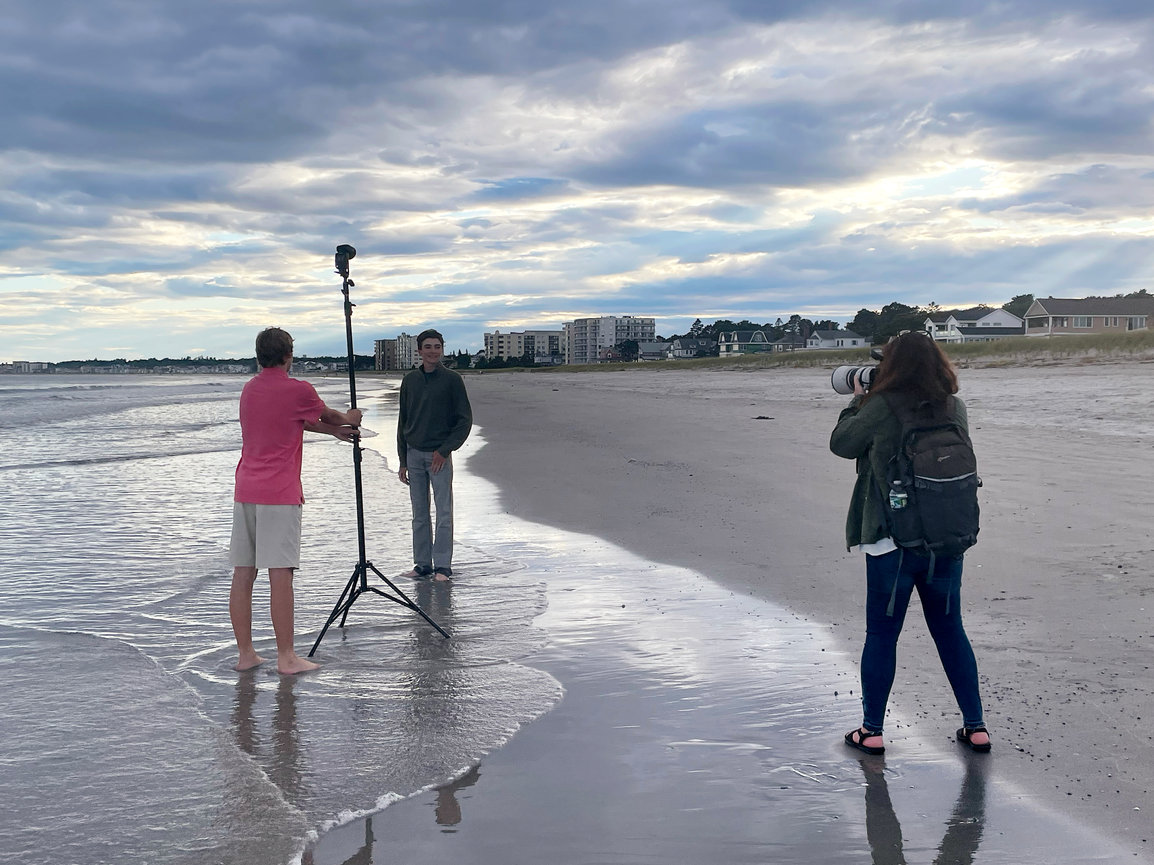 A photoshoot on at old orchard beach with a senior posing, a photographer, and a light stand. Cloudy sky in the background.