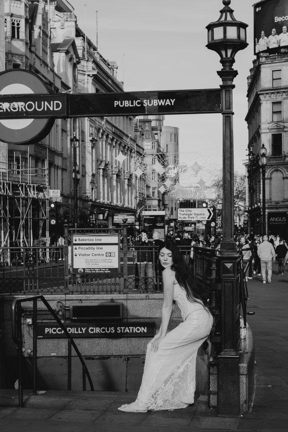 Urban London Wedding Portrait: Woman in a lace wedding dress sitting near Piccadilly Circus Station entrance in an urban street scene captured in B&W..