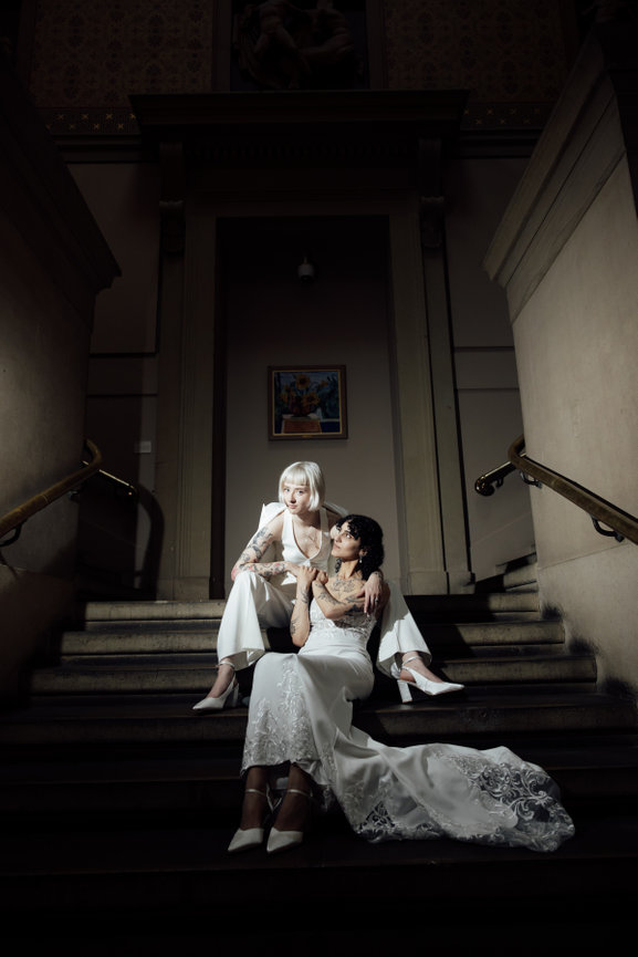 Editorial wedding photography of two brides on a grand staircase at Manchester Art Gallery, captured with direct flash for a high-fashion, modern feel.