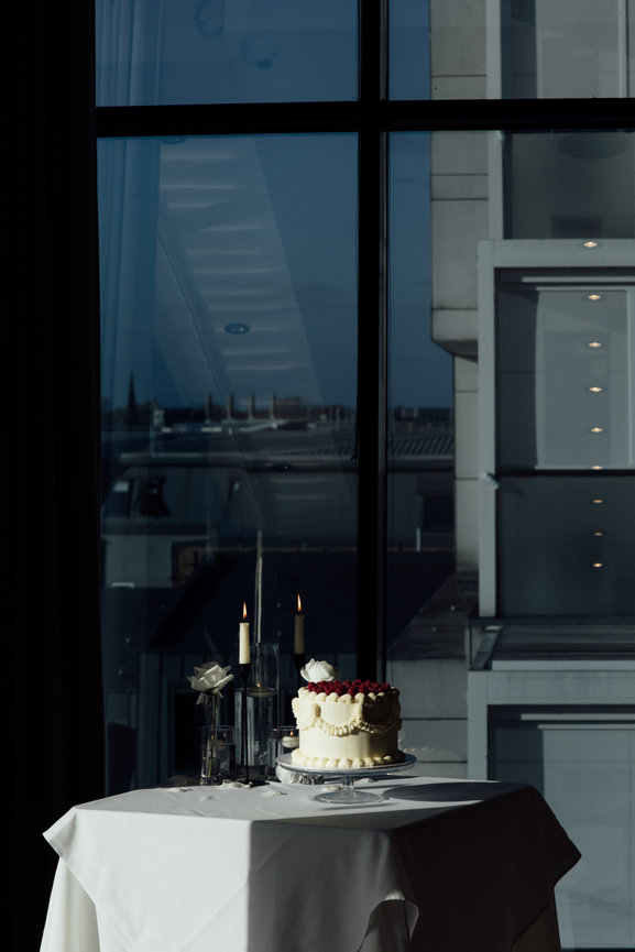 Editorial Photography at Hope Street Hotel: A wedding cake topped with raspberries on a table, beside lit candles, and white roses, set against a backdrop of Liverpool cityscape through a window.