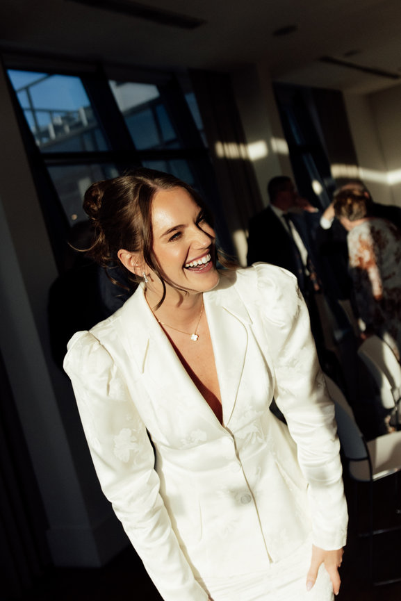 Cinematic wedding portrait of a bride in a white jacquard jacket laughing in golden hour indoor sunlight, captured with a modern editorial feel.