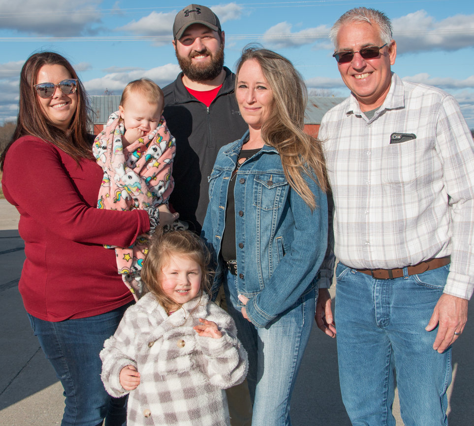 A family stands together outdoors, smiling, with a baby and young child in front.