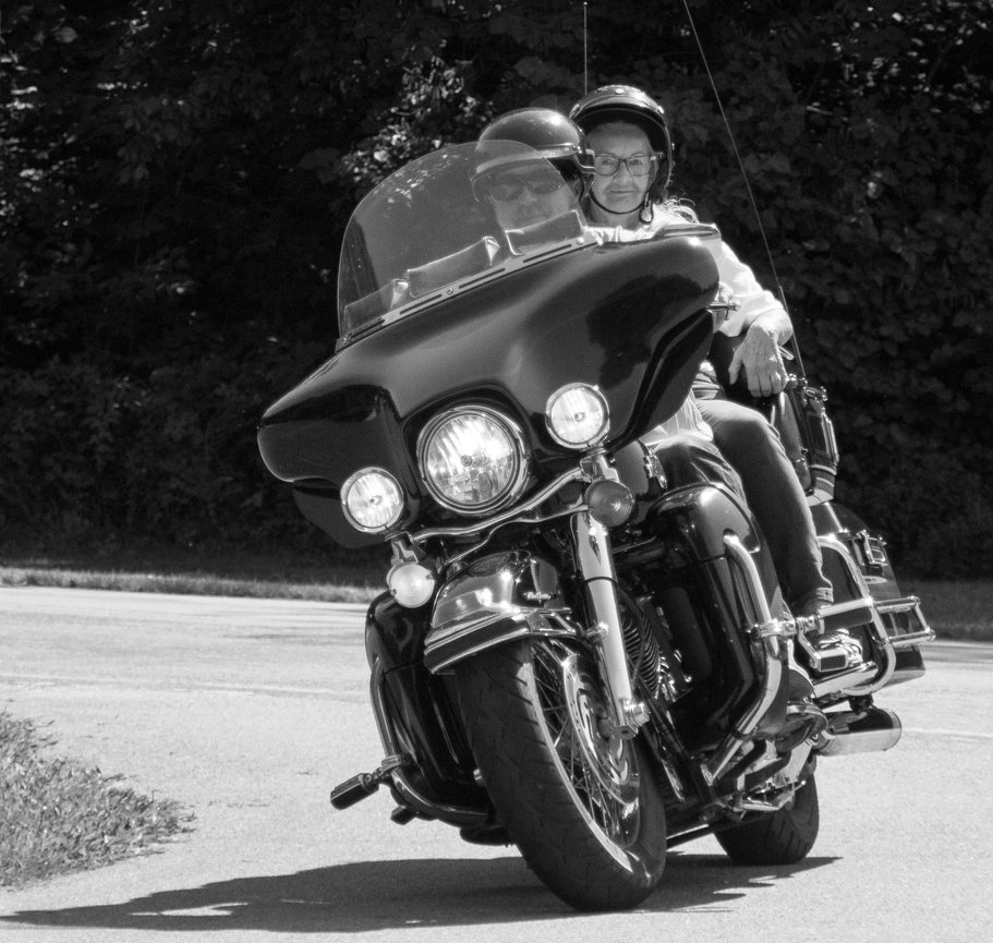 Two people ride a large motorcycle on a road, both wearing helmets, with trees in the background, in black and white.
