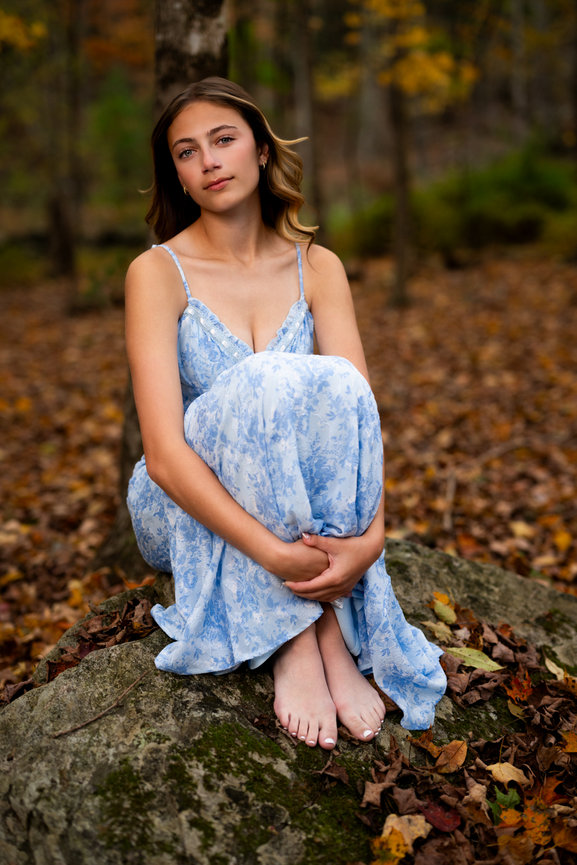 A high school senior in a blue dress sits on a rock in a forest with autumn leaves in Dallas Pennsylvania, photographed by Darren Elias Photography.